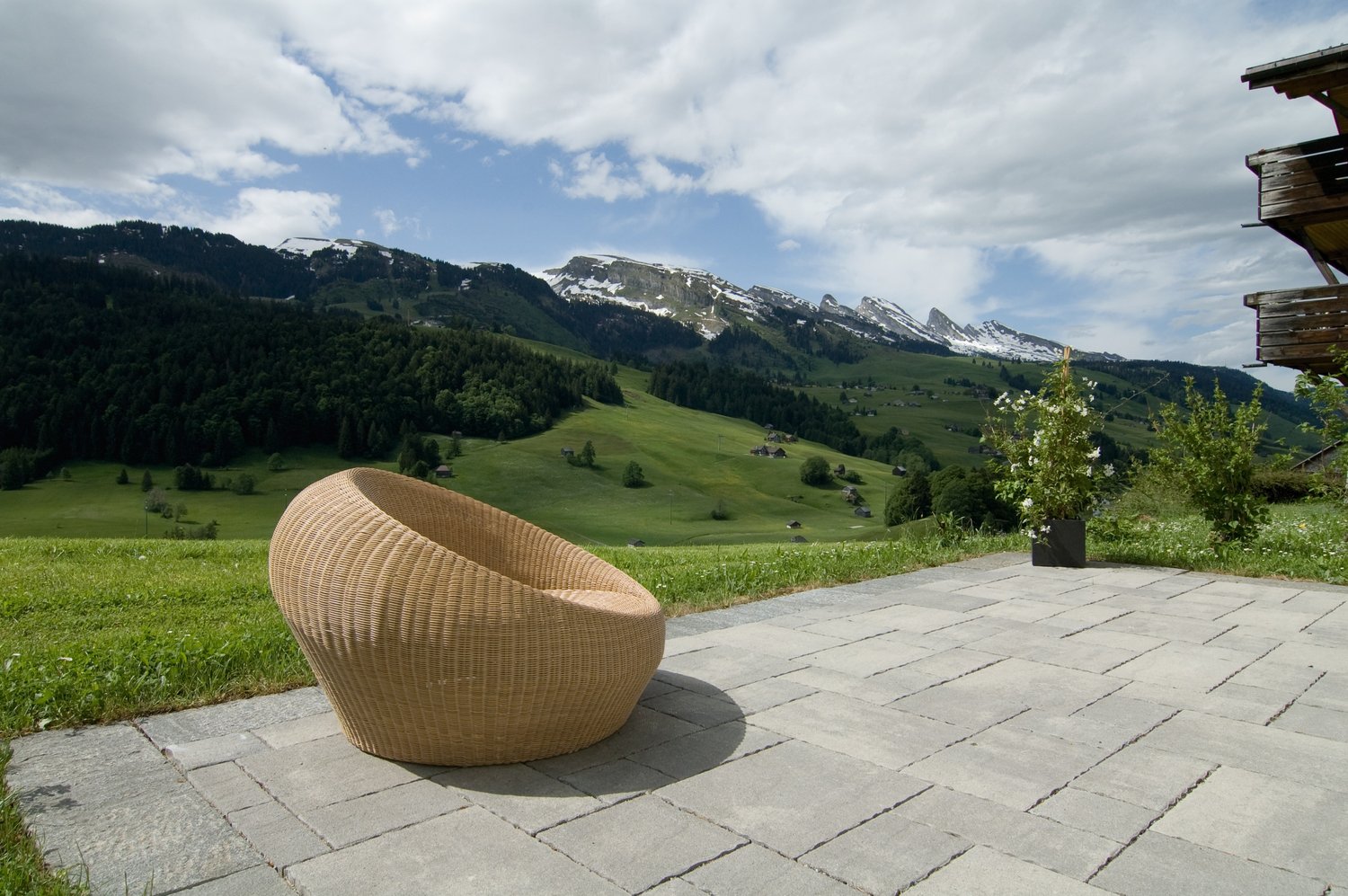 The image shows an outdoor patio area with a wicker chair overlooking a scenic mountain landscape. The patio is made of stone tiles and there is a small potted plant nearby. In the background, there are snow-capped mountains and a forested valley with gre