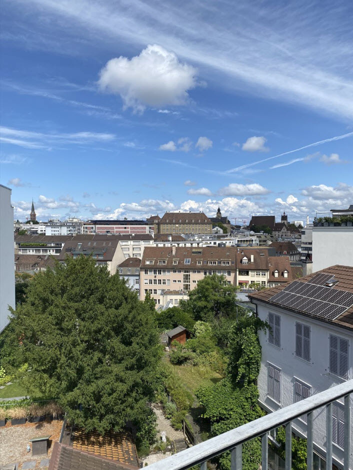 The image shows a panoramic view of a city skyline. The scene is dominated by a blue sky with fluffy white clouds. The cityscape is composed of a mix of residential and commercial buildings, with some greenery visible in the foreground. The buildings have