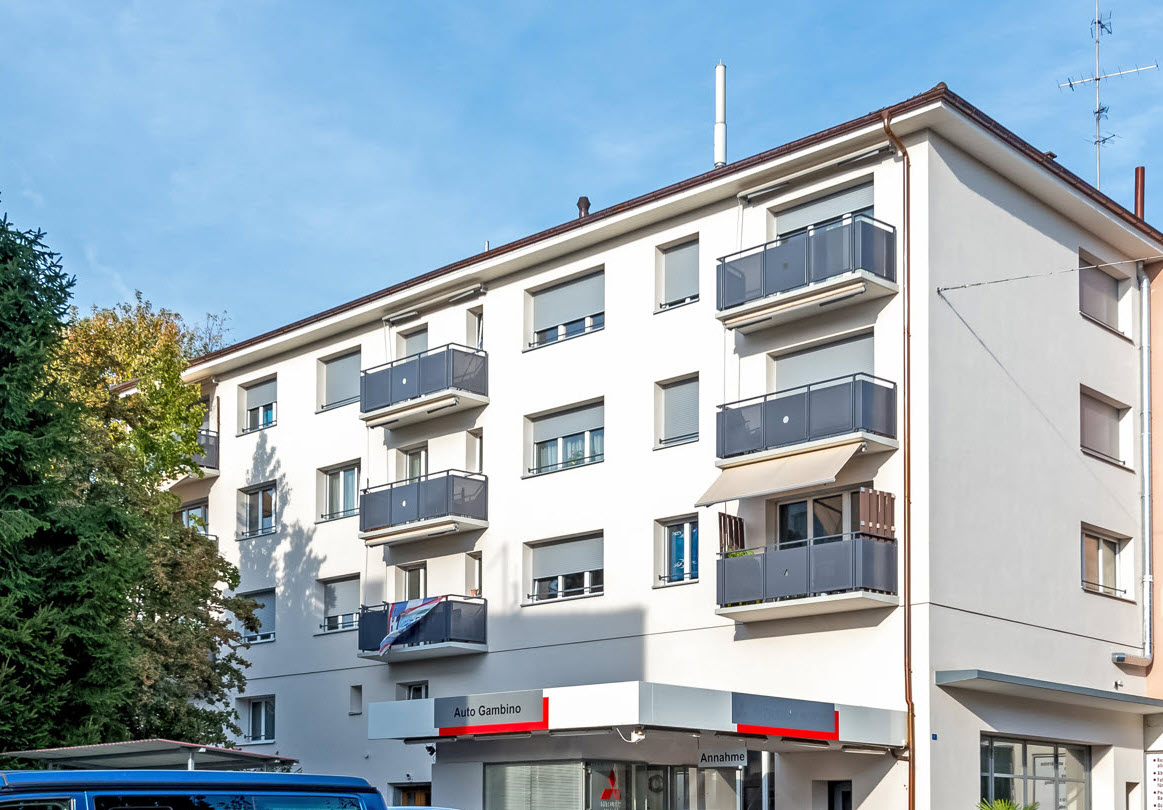 The image shows a multi-story apartment building with several balconies visible on the facade. The building has a modern, clean design with white walls and gray balcony railings. There is a commercial space on the ground floor, with the sign 'Auto Gambino