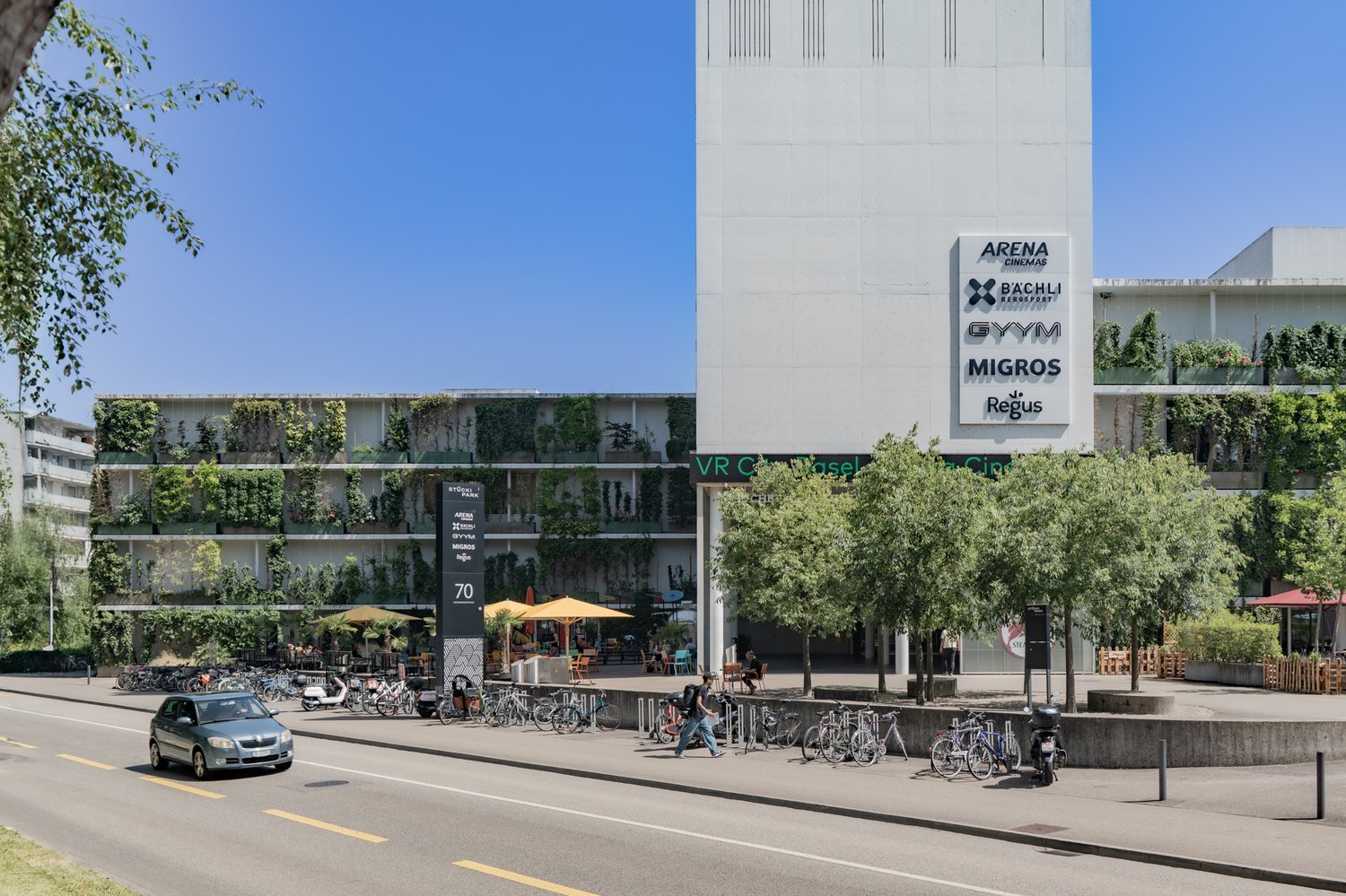 Multi-story building, vertical garden, parking area, street view, outdoor seating area, bikes parked on the side, car driving, trees and greenery, multiple shops on the ground floor, white walls and signs.