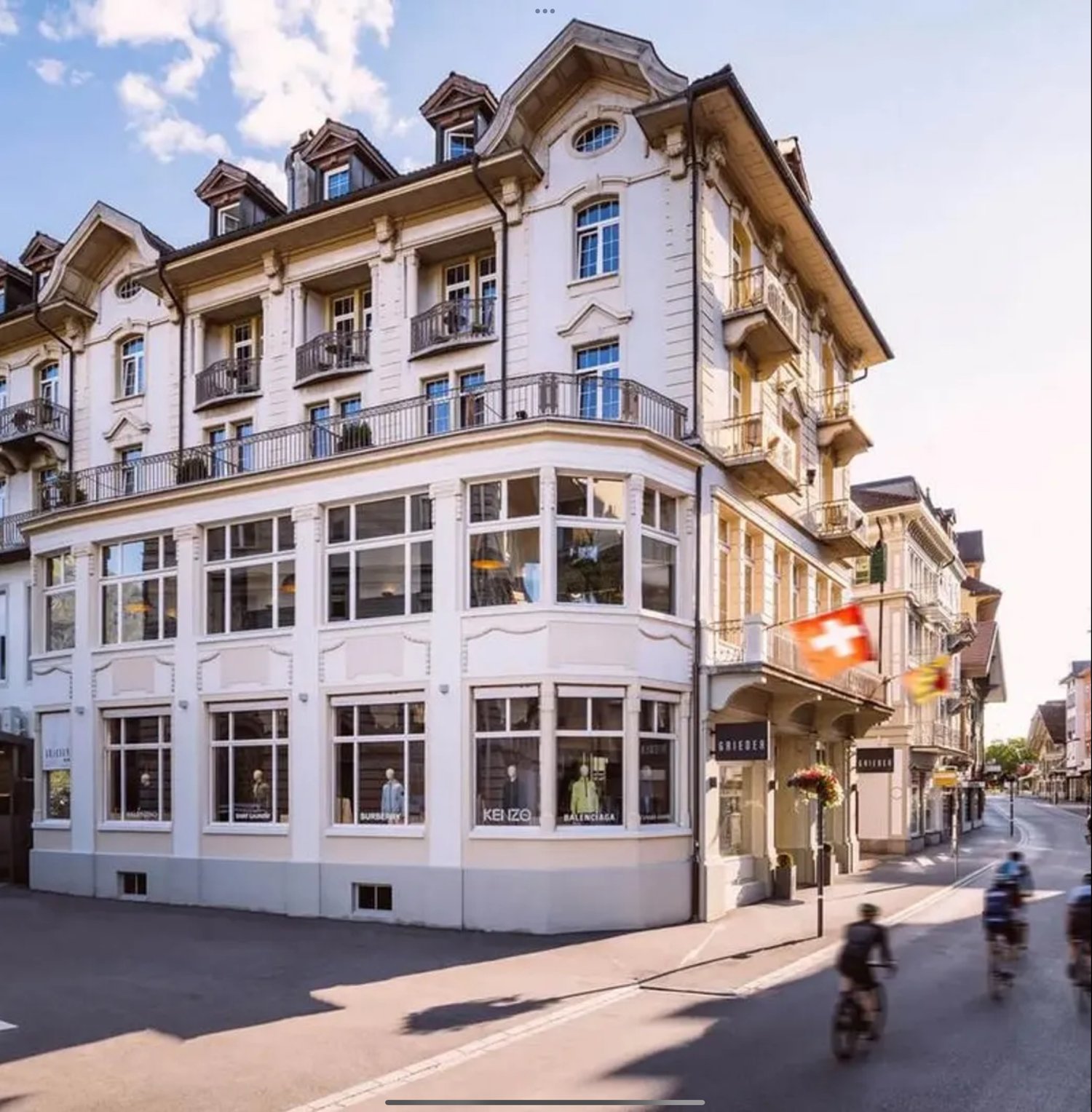 The building is a multistory white building with large windows. It has several floors and a lot of windows. The building looks modern and elegant with no visible exterior balconies. It has a balcony at the top floor. The building also features a triangula