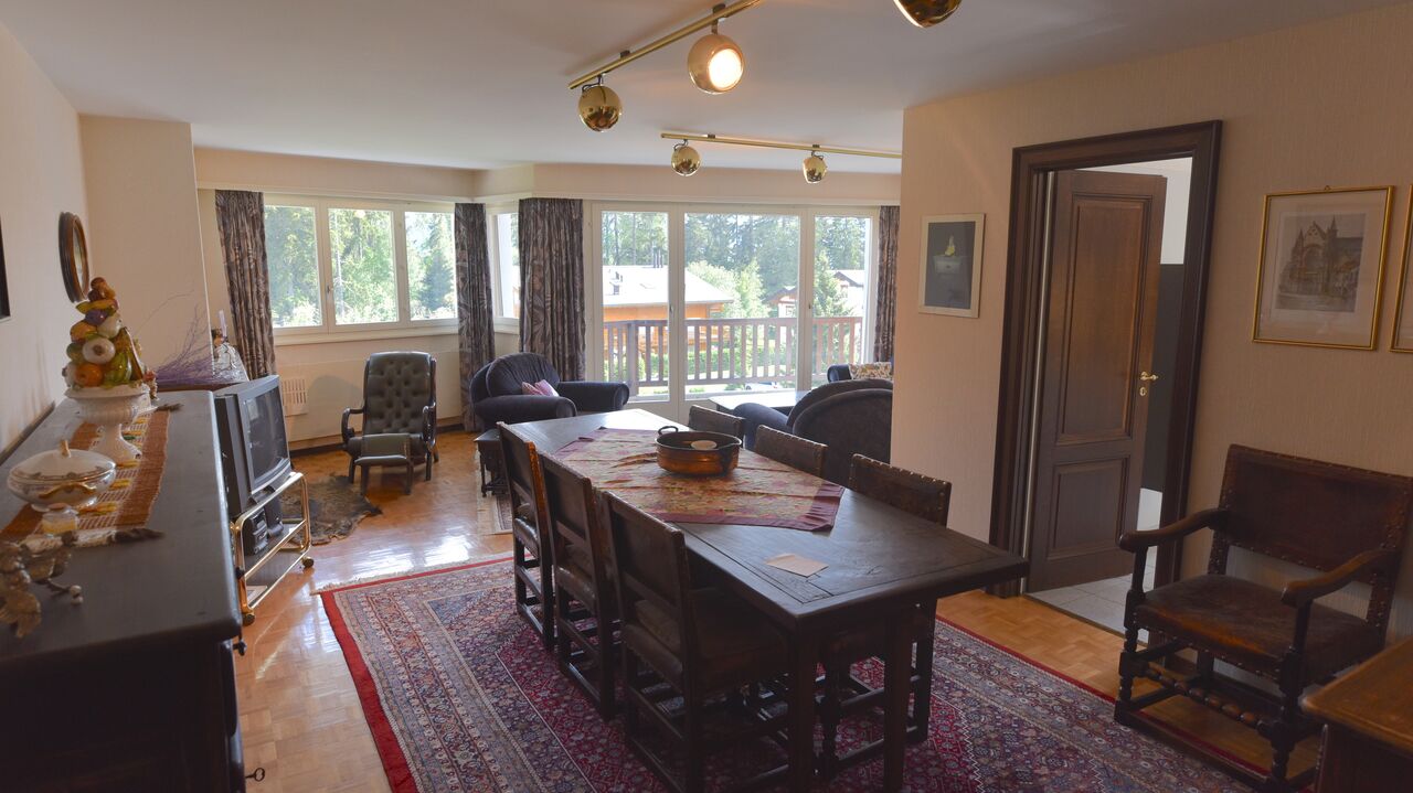 The image shows a spacious open-plan living and dining area. The room has large windows that provide a view of a balcony or terrace overlooking a wooded area. The living room features comfortable sofas and chairs, while the dining area has a wooden table