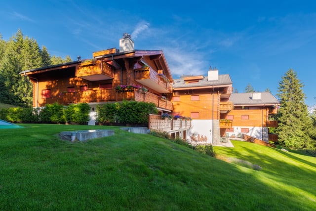 2-story wooden chalet-style house with balconies, surrounded by lush green lawn and trees, set against a blue sky