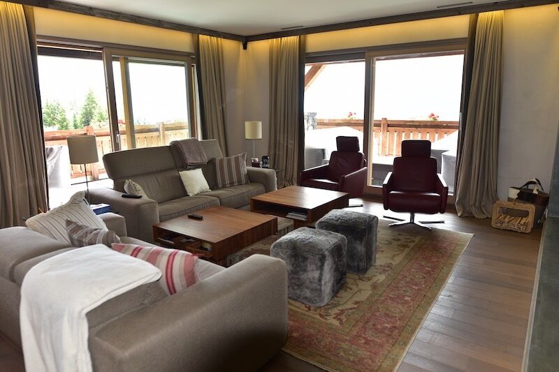 Spacious living room with large windows, hardwood floors, comfortable sofas and armchairs, wooden coffee table, and a balcony or terrace visible through the windows.