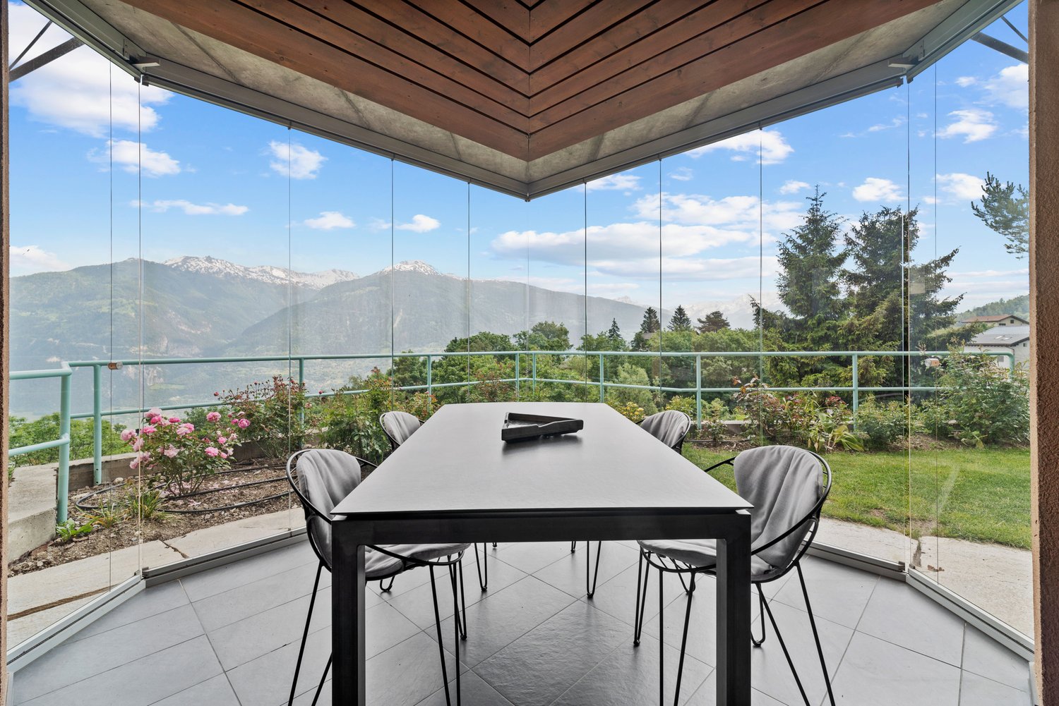 Spacious dining area with a large glass-enclosed patio, offering a panoramic view of the surrounding mountains and lush greenery. The room features a modern black dining table and chairs, with a wooden ceiling and beams providing a warm, rustic ambiance.