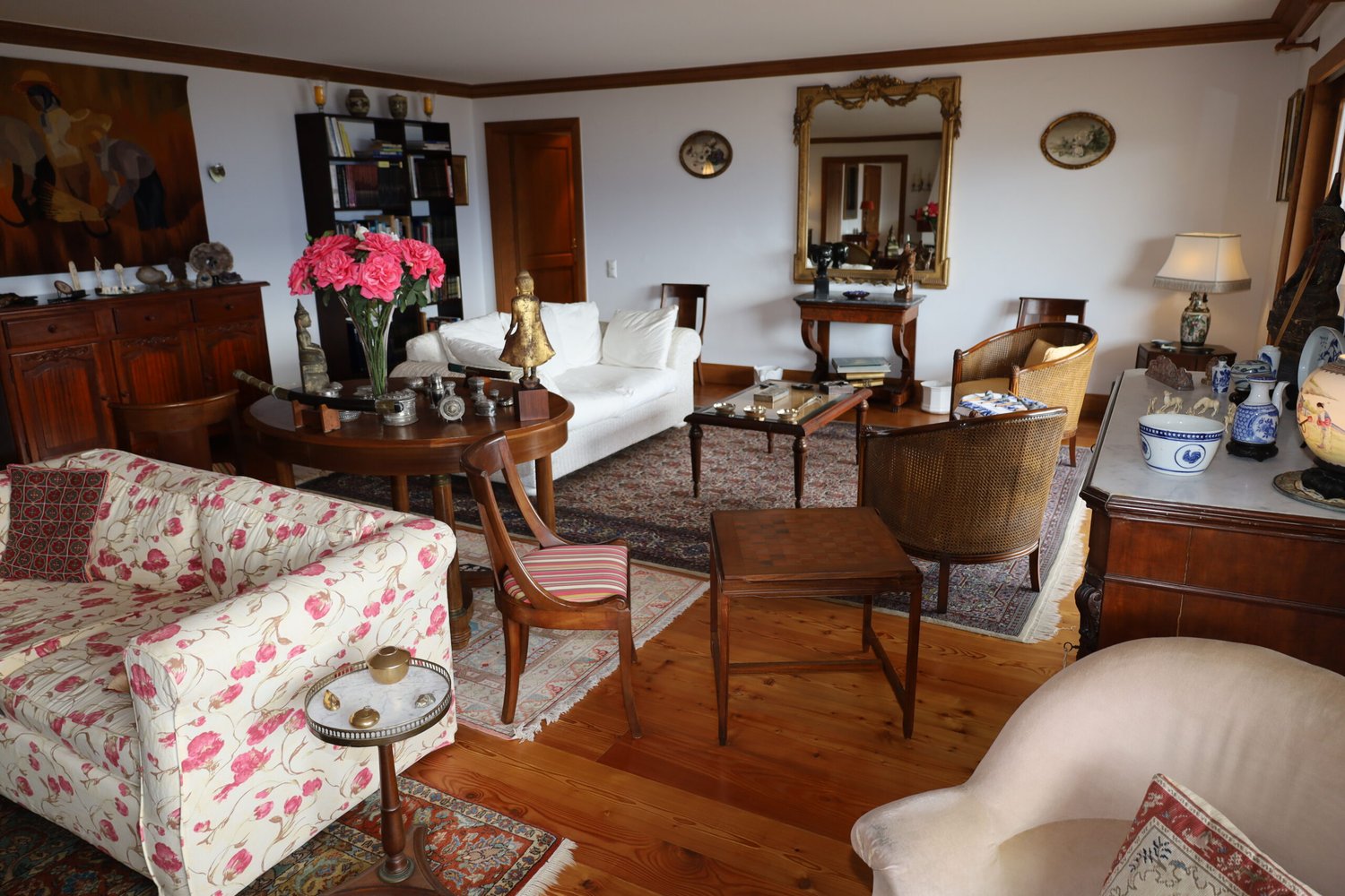Spacious living room with a mix of traditional and vintage furniture, including a floral patterned sofa, wooden chairs, a coffee table, and a bookshelf. The room features hardwood floors, white walls, and decorative elements such as a large mirror, artwor