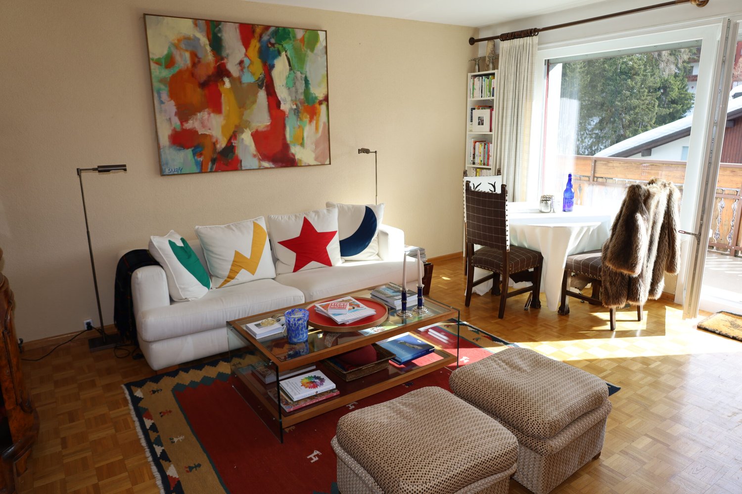 living room: white couch with colorful pillows, glass coffee table, wooden floor, abstract painting on wall, large window, balcony with wooden railing, view of mountains