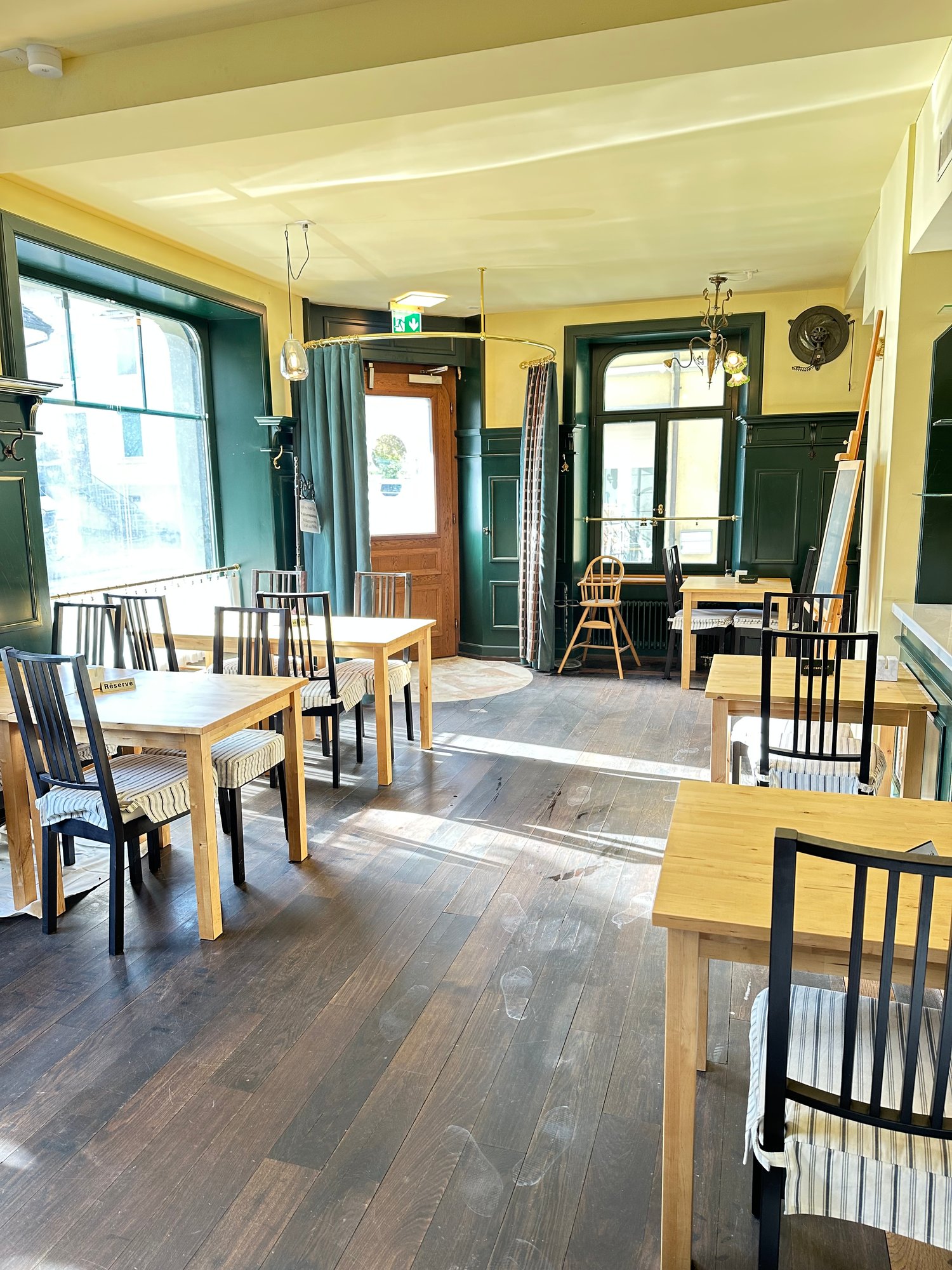 Wooden floor, rectangular dining tables, wooden chairs with striped cushions, green walls, wooden door, large windows, ceiling lamps