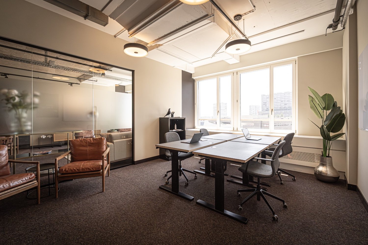 Empty office room with desks and chairs, large windows, potted plant, leather chair, small table