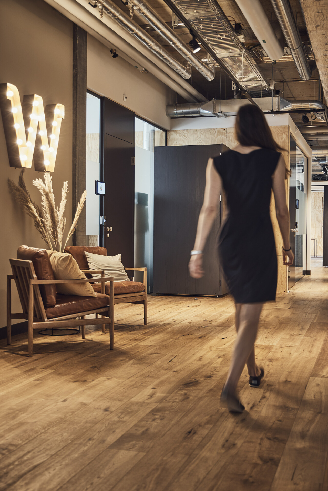 Large room with modern lighting, wooden floor, decorative pillows, a woman walking, brown leather armchairs, a 'W' light sign on the wall
