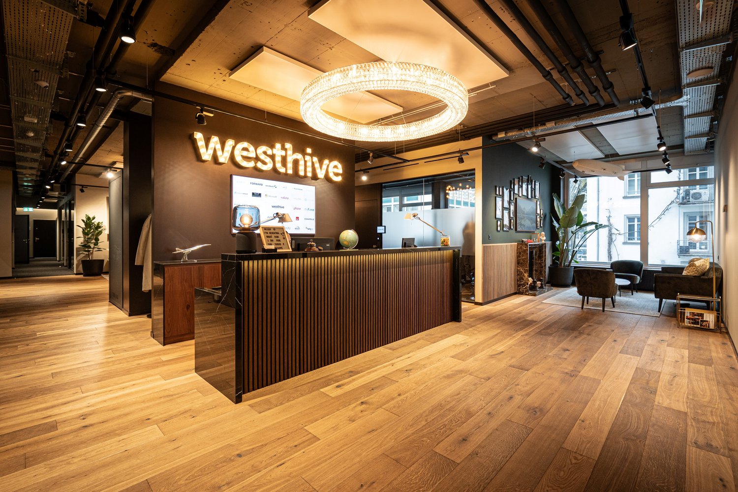 Wooden flooring, large chandelier, glass windows, wooden reception desk, couch, desk, large plant, sign