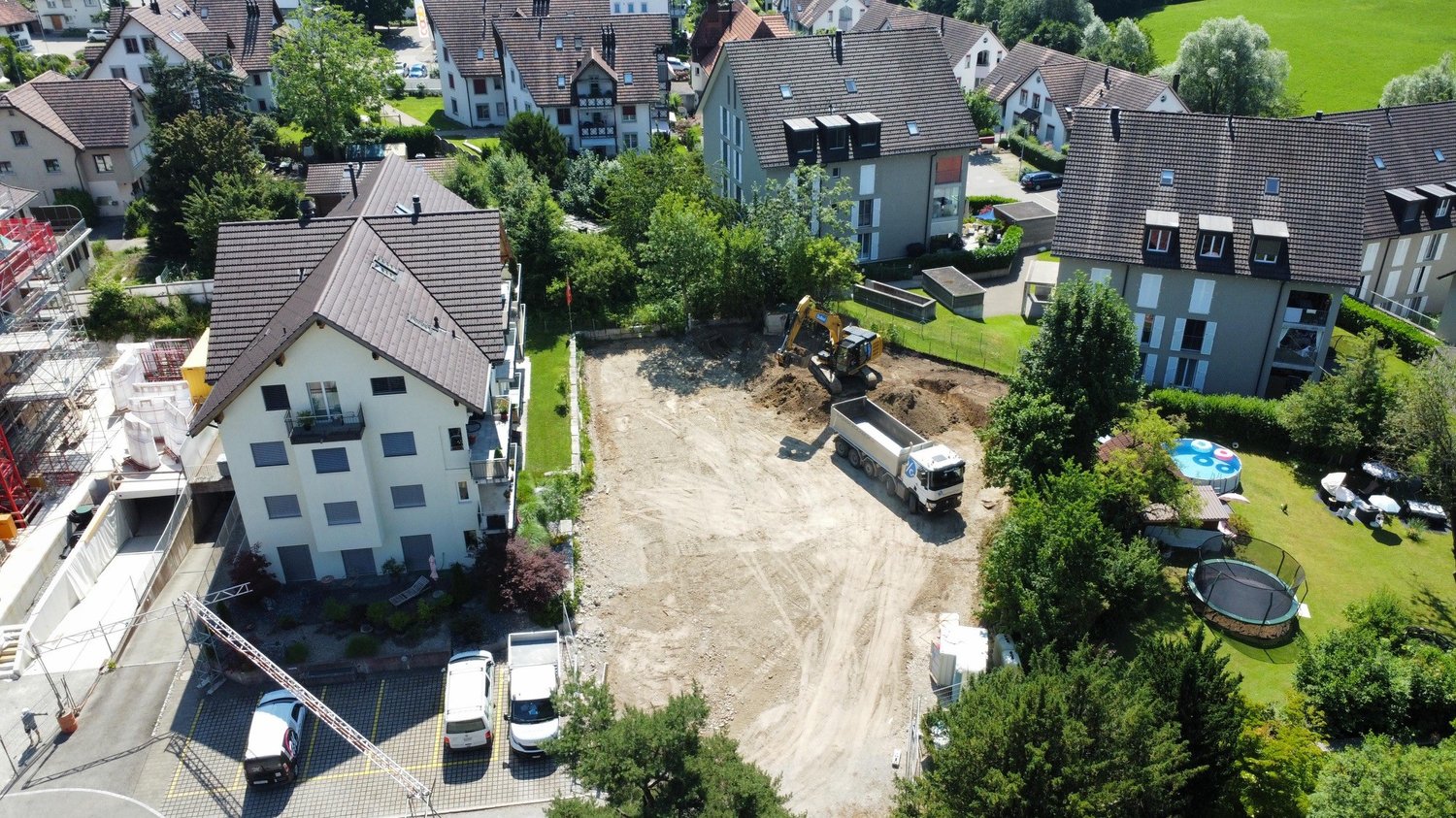 Aerial view of a residential area with a construction site in the center. The site has an excavator and a dump truck, indicating ongoing construction work. The surrounding buildings appear to be multi-story residential apartments with tiled roofs. There a