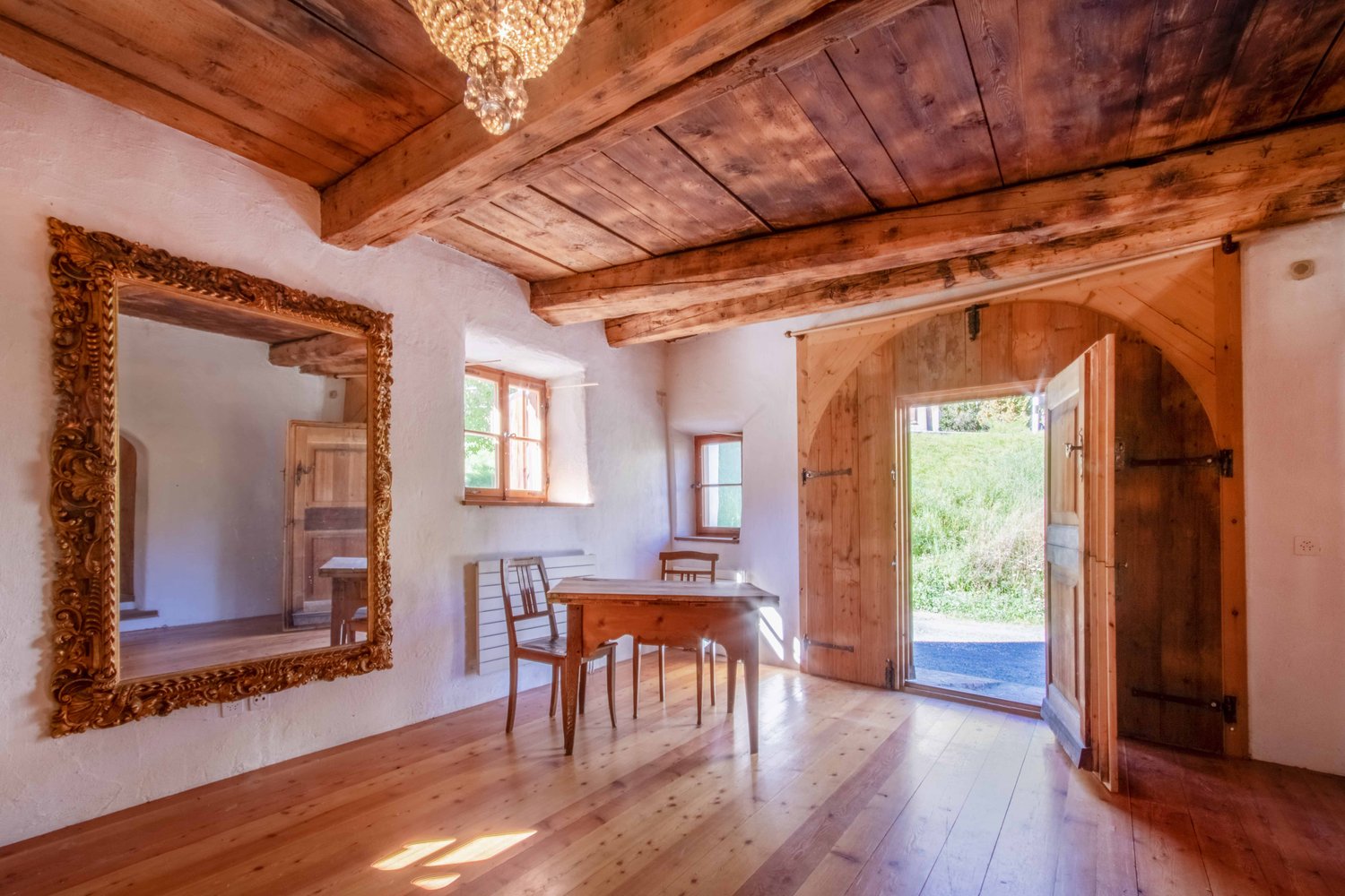 Dining room: Wooden floor, wooden dining table and chairs, ornate mirror, wooden ceiling with chandelier, wooden door, window with white shutter