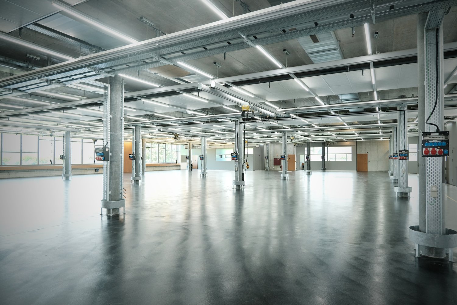 Empty warehouse with metal beams, exposed pipes, large windows, concrete floor.
