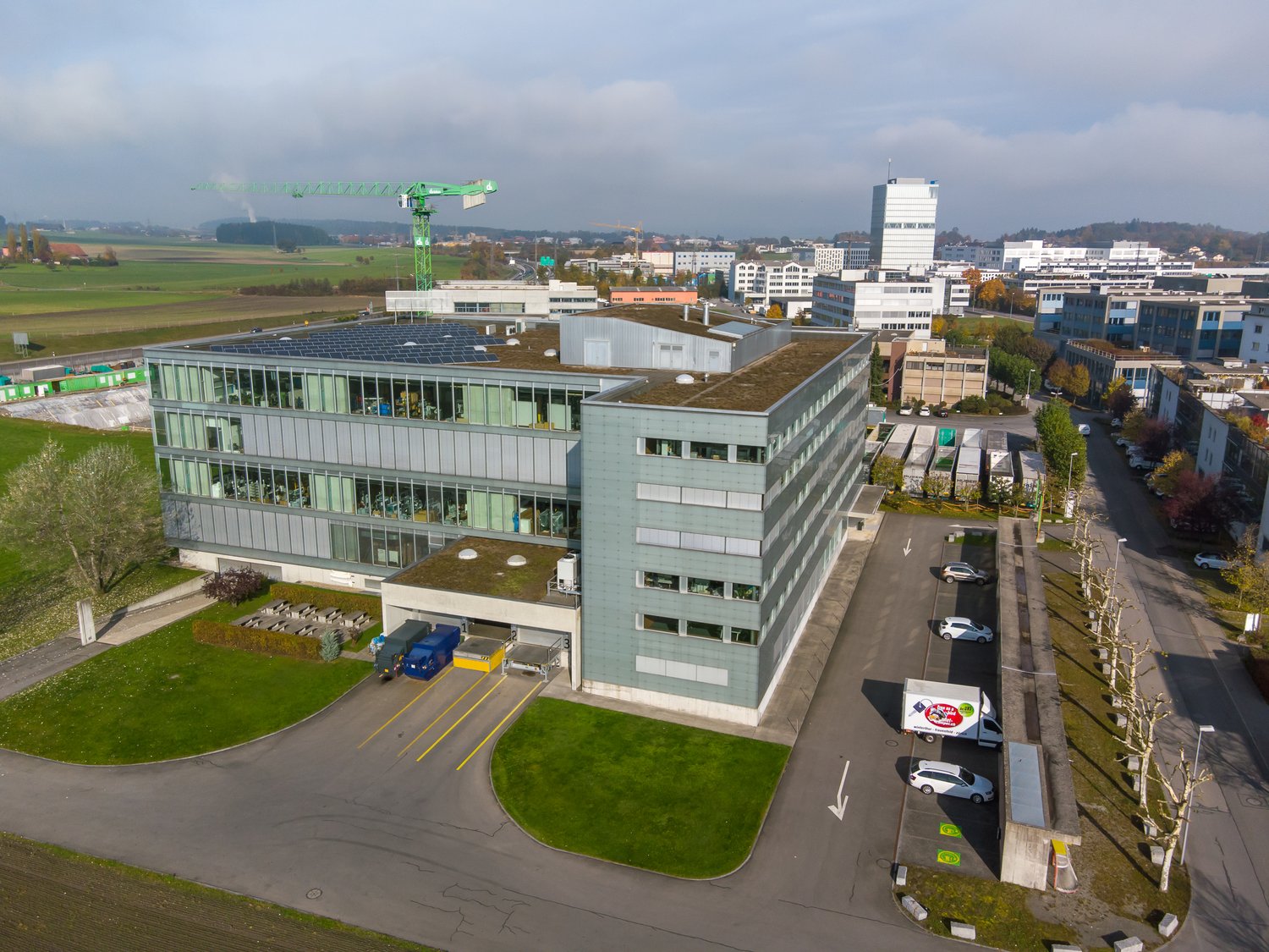 Modern office building with solar panels on the roof, multiple glass windows, several vehicles parked, landscaped area with grass and trees