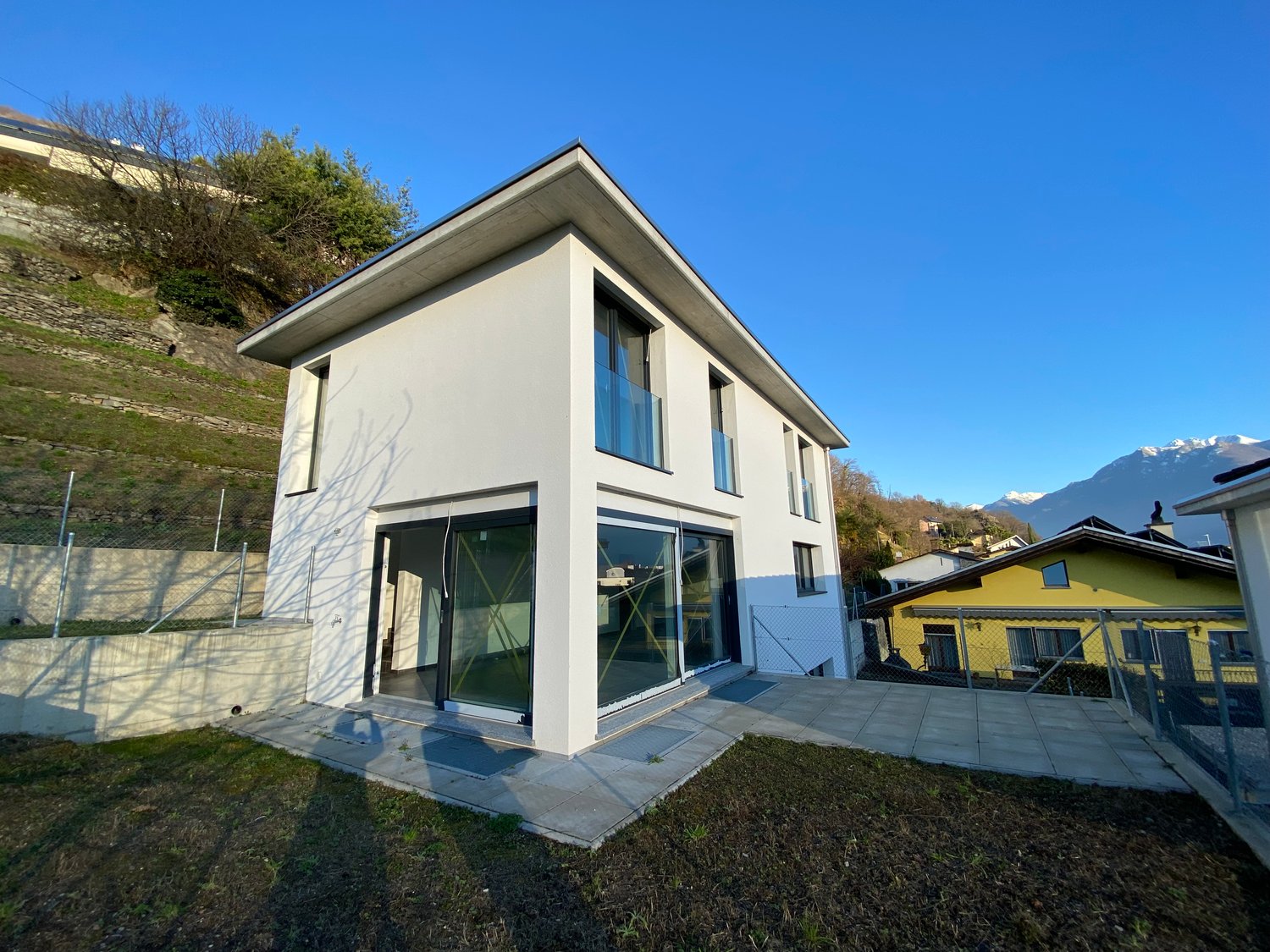 Modern white house, large glass windows, modern architecture, sloping ground with vegetation, small porch with glass doors, concrete fence, mountains in the background.