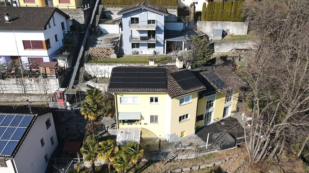 Two-story yellow house with solar panels on the roof, surrounded by other buildings, in a neighborhood with trees.