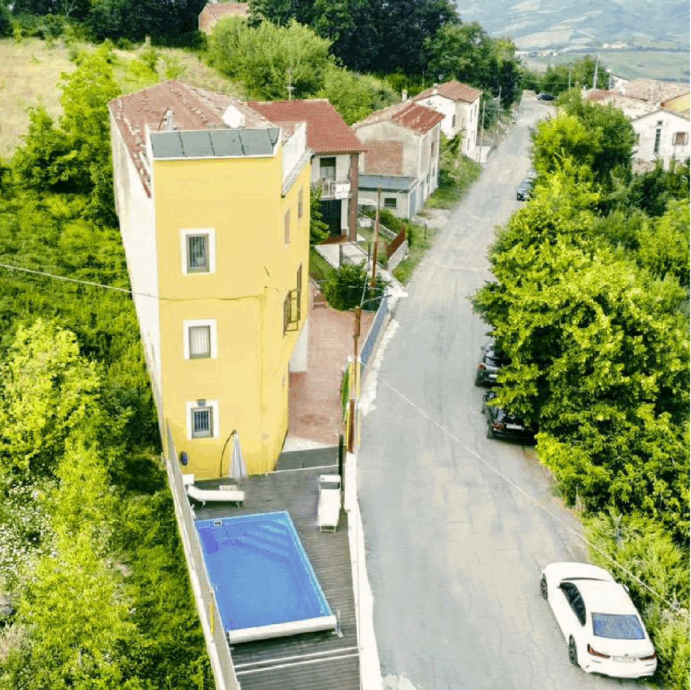 Building with yellow walls, swimming pool, red roofs, surrounded by houses and vegetation