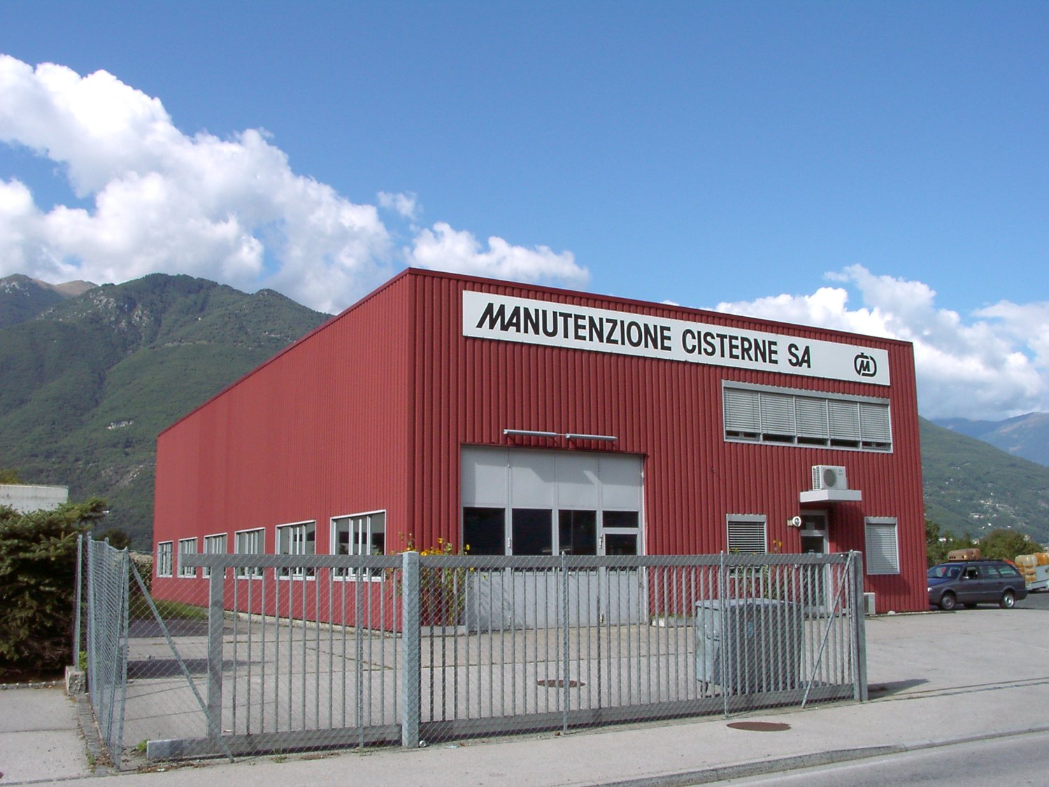 Red industrial building with large garage doors, multiple windows, and a sign reading 'MANUTENZIONE CISTERNE SA'.