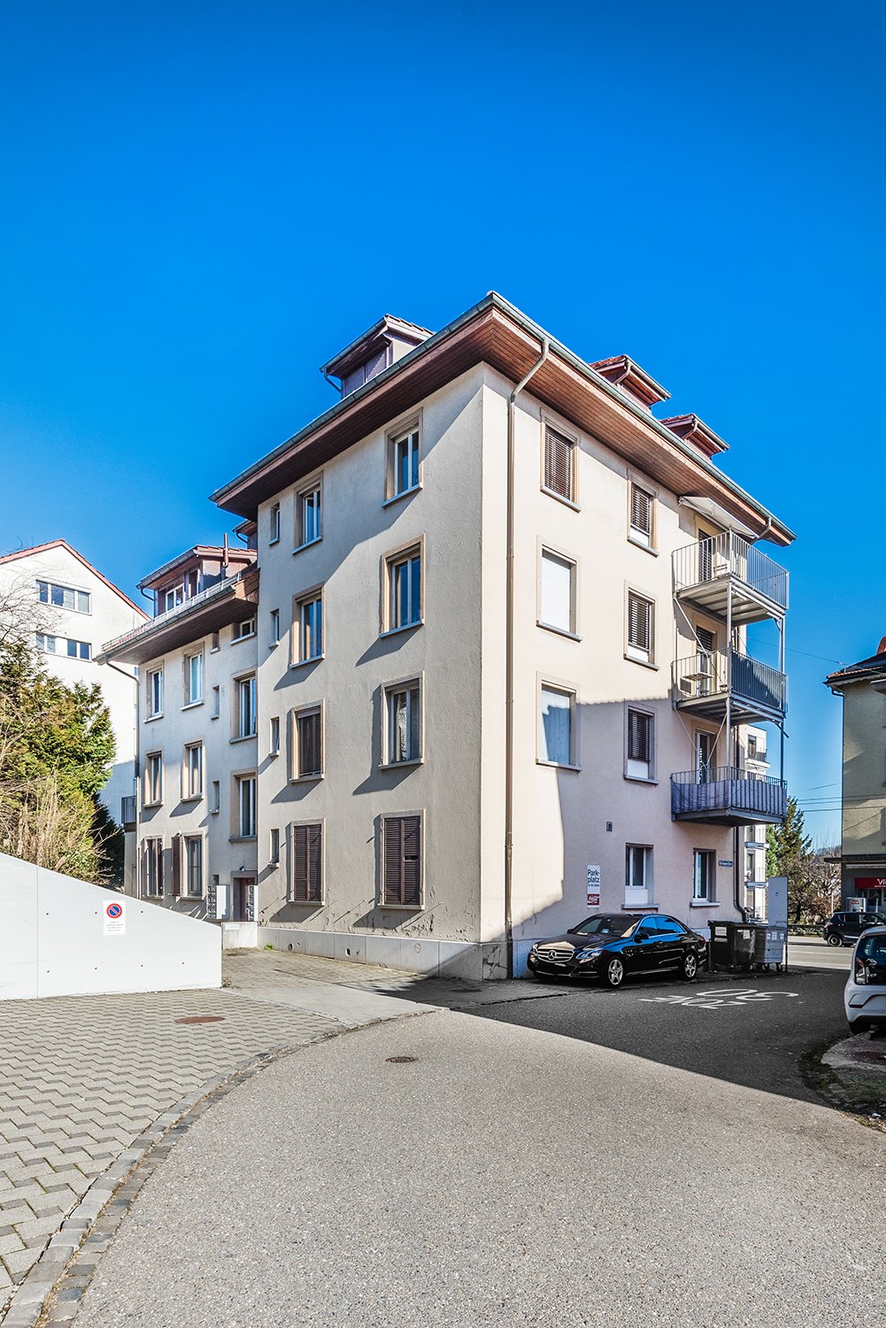 White building with multiple windows, balconies, a car parked in front, trash bins on the side, and another car parked nearby.