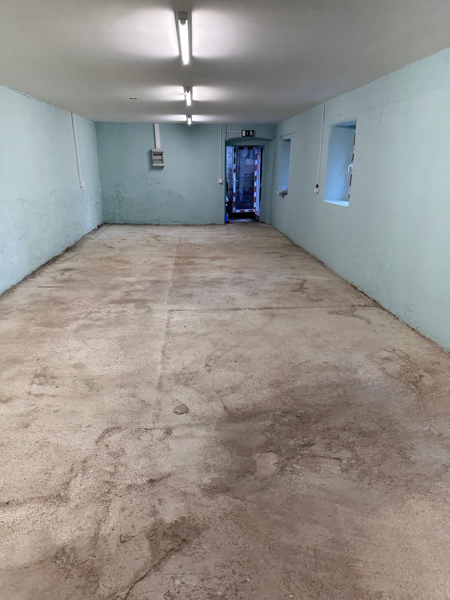 Empty room with light grey walls and concrete floor. There are fluorescent ceiling lights. There are small windows, one of which has a door.