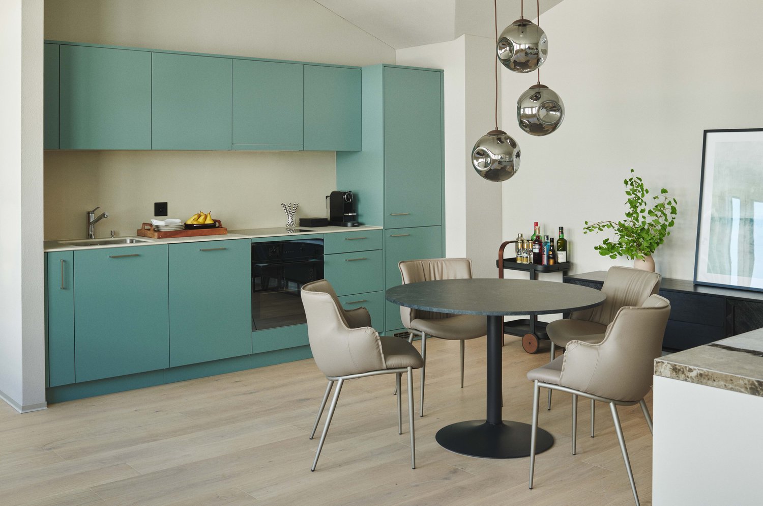 The image shows a modern kitchen and dining area. The kitchen features teal-colored cabinets and a built-in dishwasher. The dining area has a round table with beige chairs. The room is illuminated by pendant lights and has a plant decoration. The overall