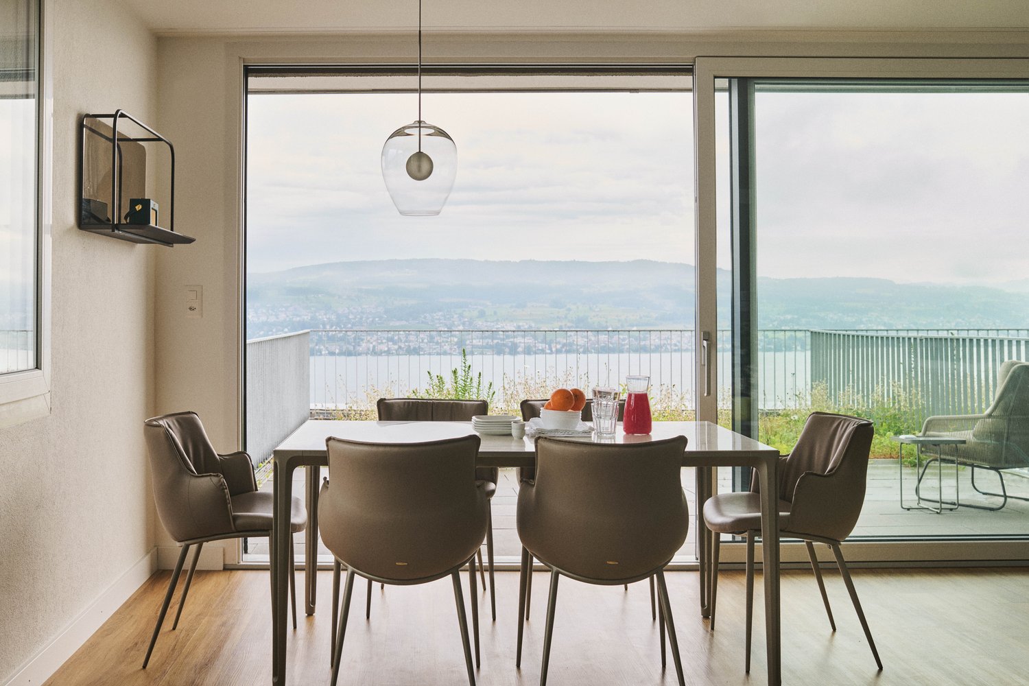 modern dining room with a glass table, modern chairs, glass wall and balcony with a view over a lake and mountains