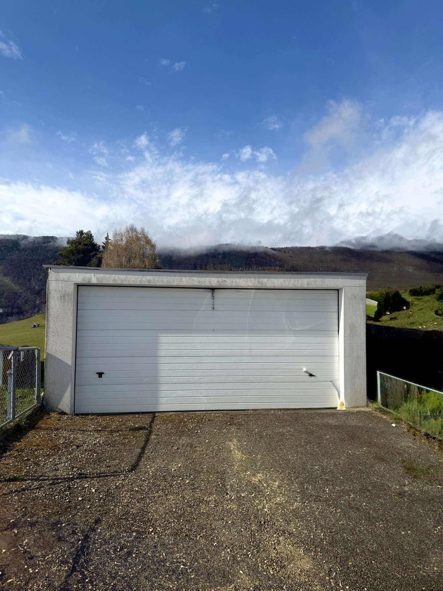 Garage with a white door, concrete walls, gravel driveway, fenced area, green field, mountains in the background