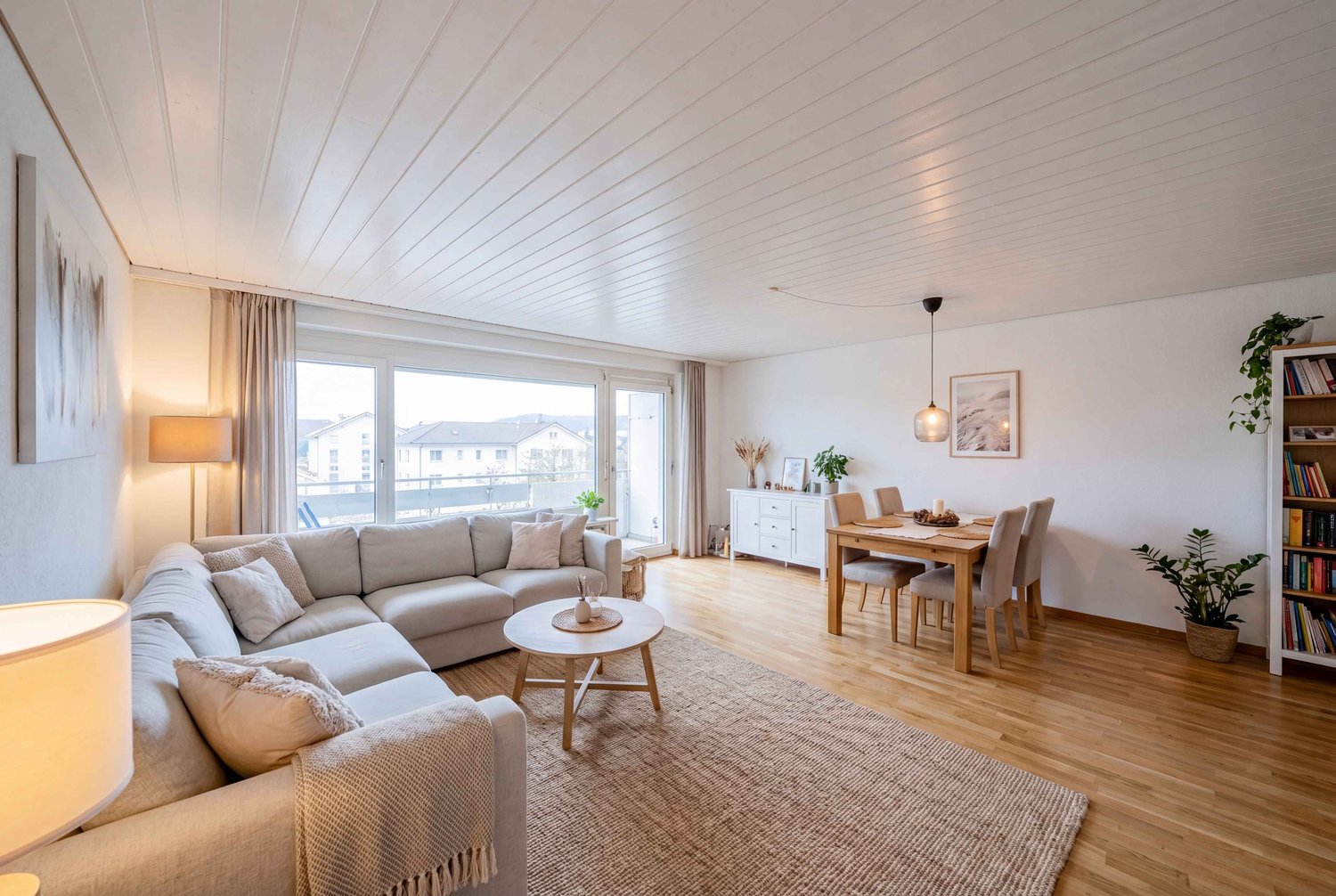 large windows, gray sofa, wooden coffee table, wooden dining table, wooden floor, white walls, ceiling lamp