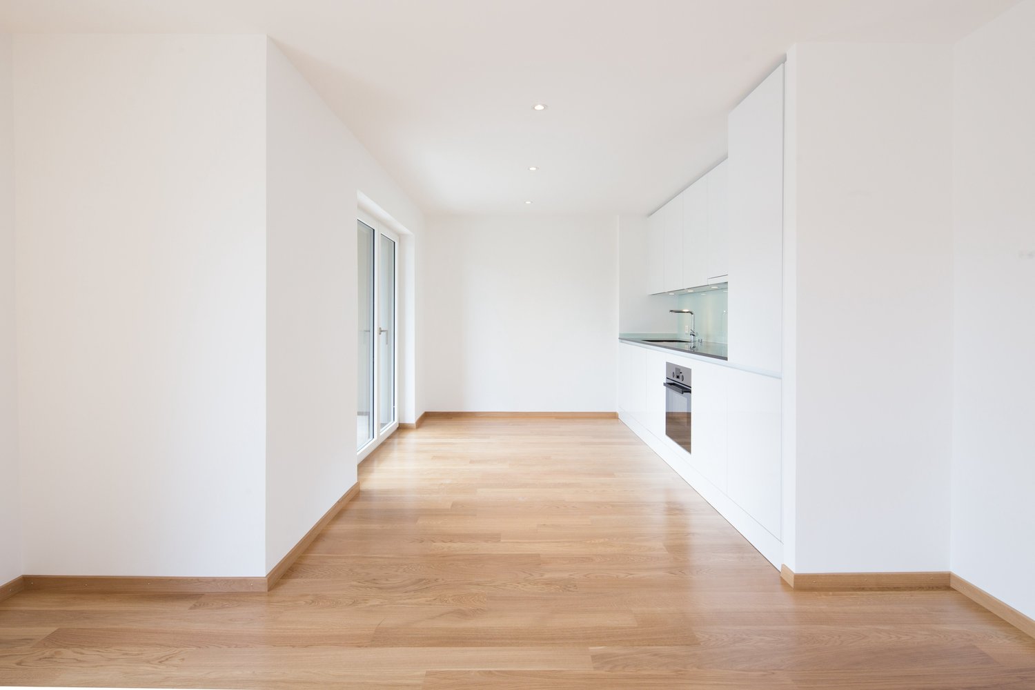 Empty room with wooden floor, white walls, large sliding glass doors, open kitchen, sink, oven.