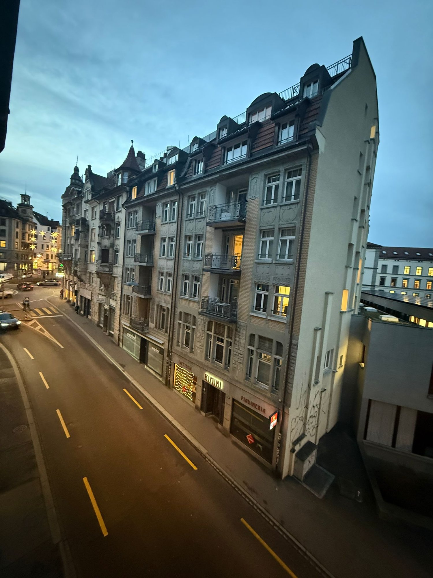 Urban apartment building, facade made of brick, glass windows, multiple floors, balconies, storefronts on the ground floor, traffic lanes, some parked vehicles, and street lamps on the sides.