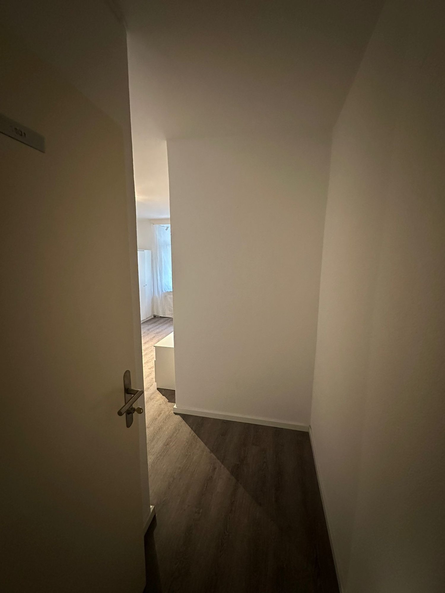 empty room, white walls, wooden floor, open door leading to bedroom, small white desk, white wardrobe, glass window with white curtain