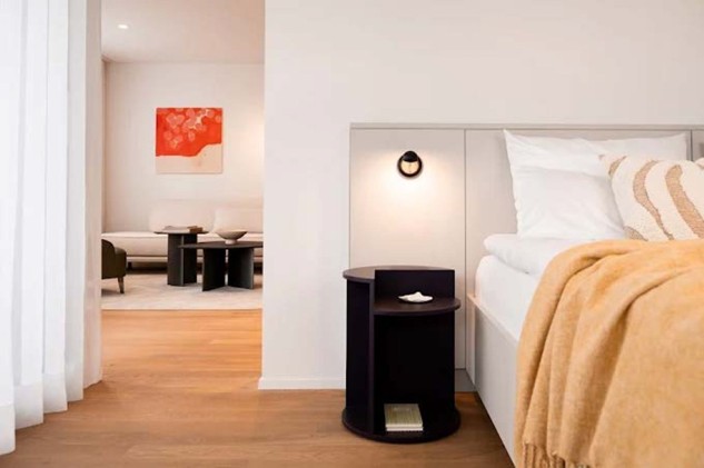 white bed with a blanket and pillows, dark night stand, lit lamp, white walls, picture on the wall, chair and table in the other room