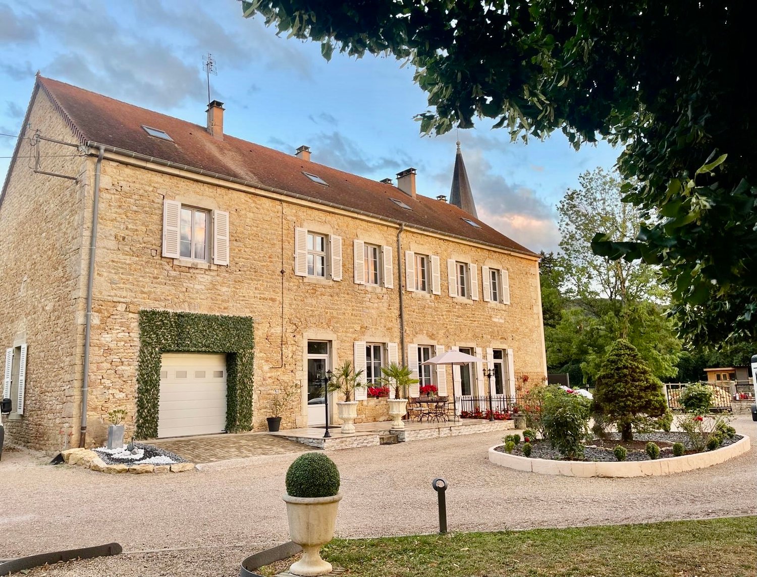 Large stone house, brown roof, white shutters, white garage, garden, driveway, outdoor seating area, plants, trees