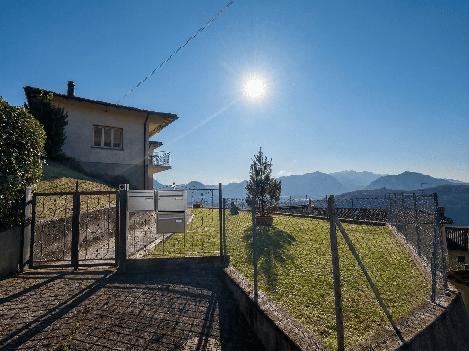 House with a balcony, surrounded by a fence, with a mailbox, a small garden, and a view of the mountains.