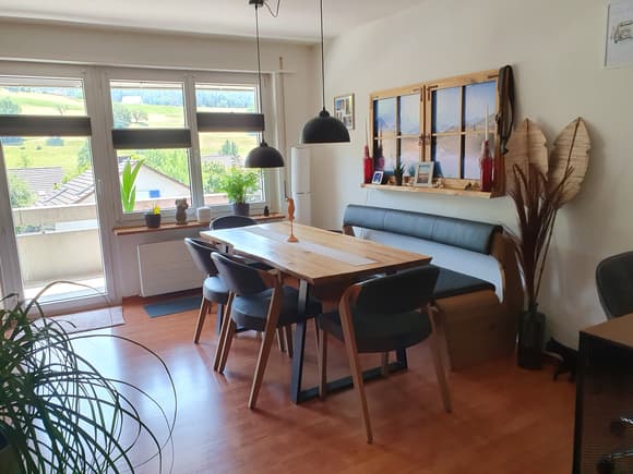 wooden dining table with 6 chairs, black bench with cushions, white wall, big windows, floor heating, plants
