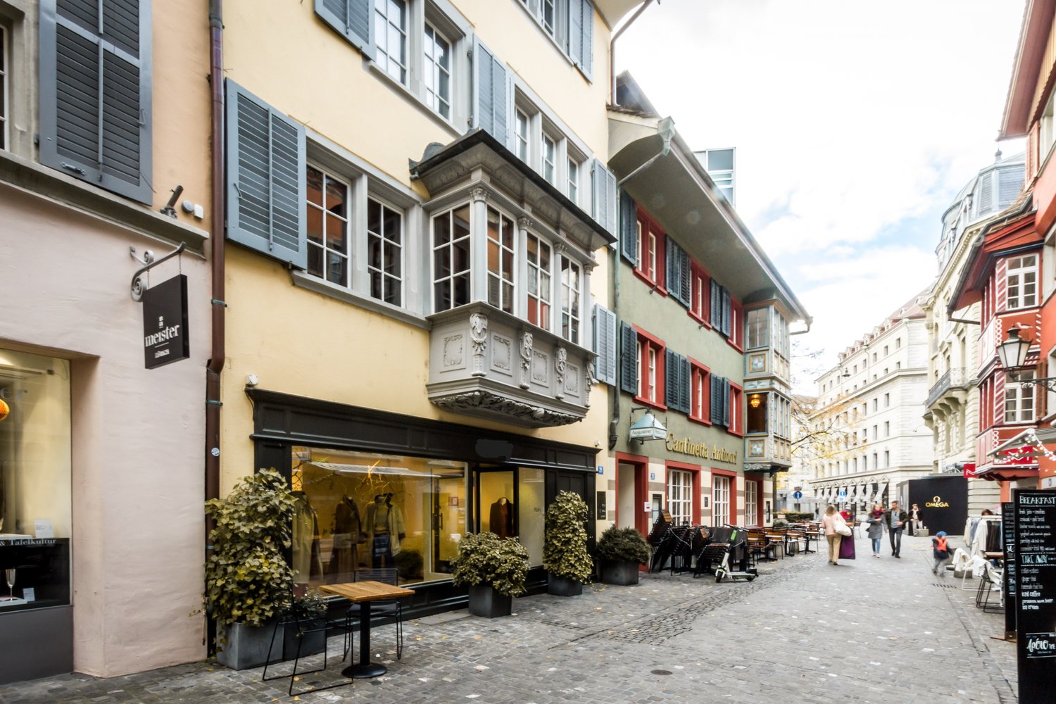 Cobblestone street, buildings with balconies, shops, people walking, outdoor seating, potted plants