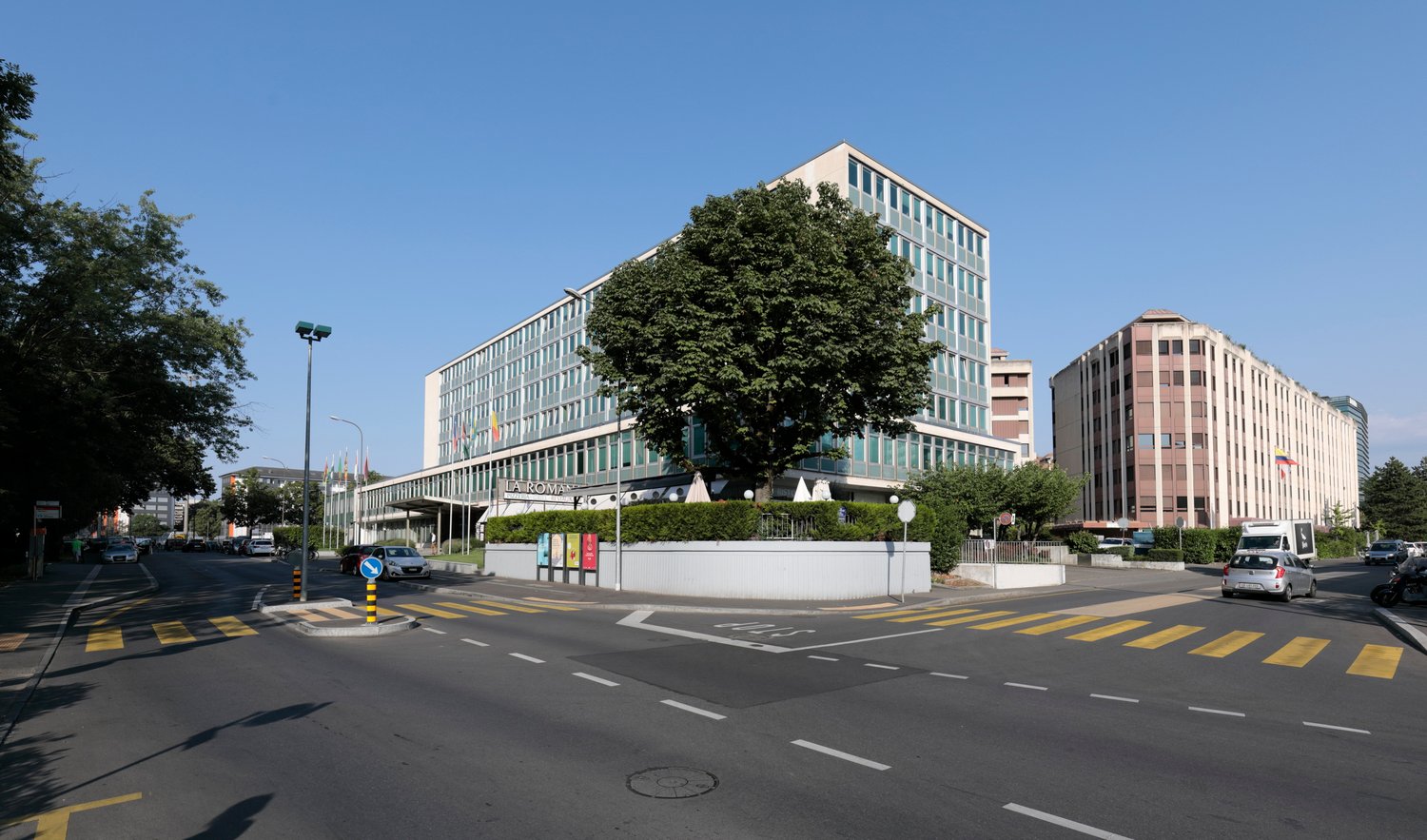 Modern office building, glass facade, multiple floors, surrounded by greenery, street with cars and pedestrians