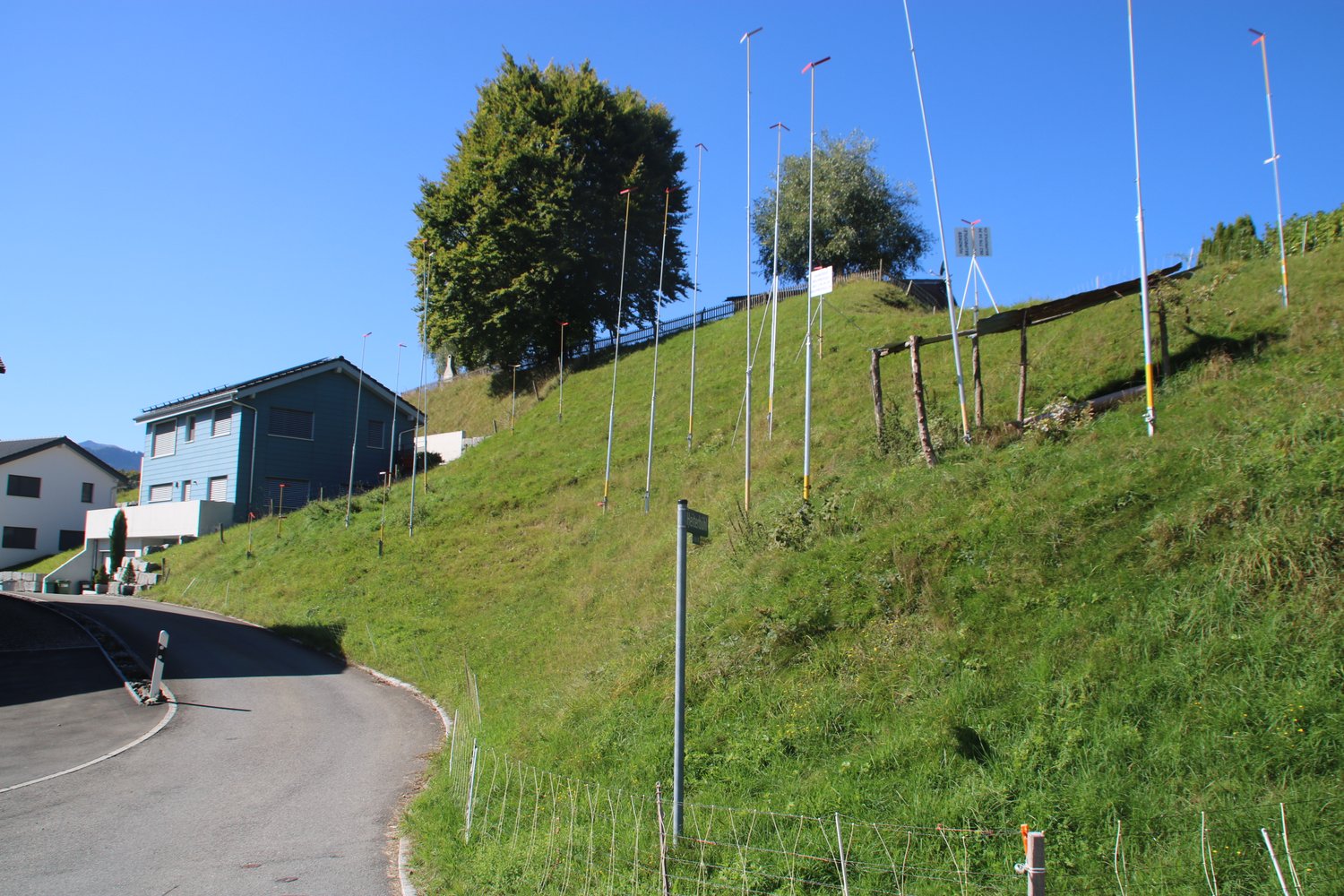 The image shows a hilly, grassy landscape with a paved road running through it. There are several buildings visible on the raised ground, including what appears to be a residential building. The landscape is dotted with trees and there are several tall po