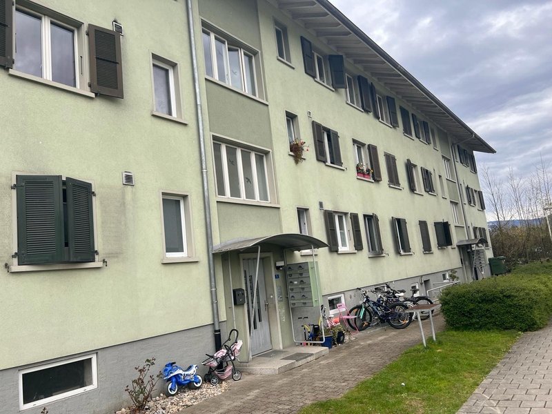 An apartment building with green walls, brown windows, and black shutters. The entrance of the building has a covered area with mailboxes. Outside, there are parked bicycles, a stroller, and a trash bin.