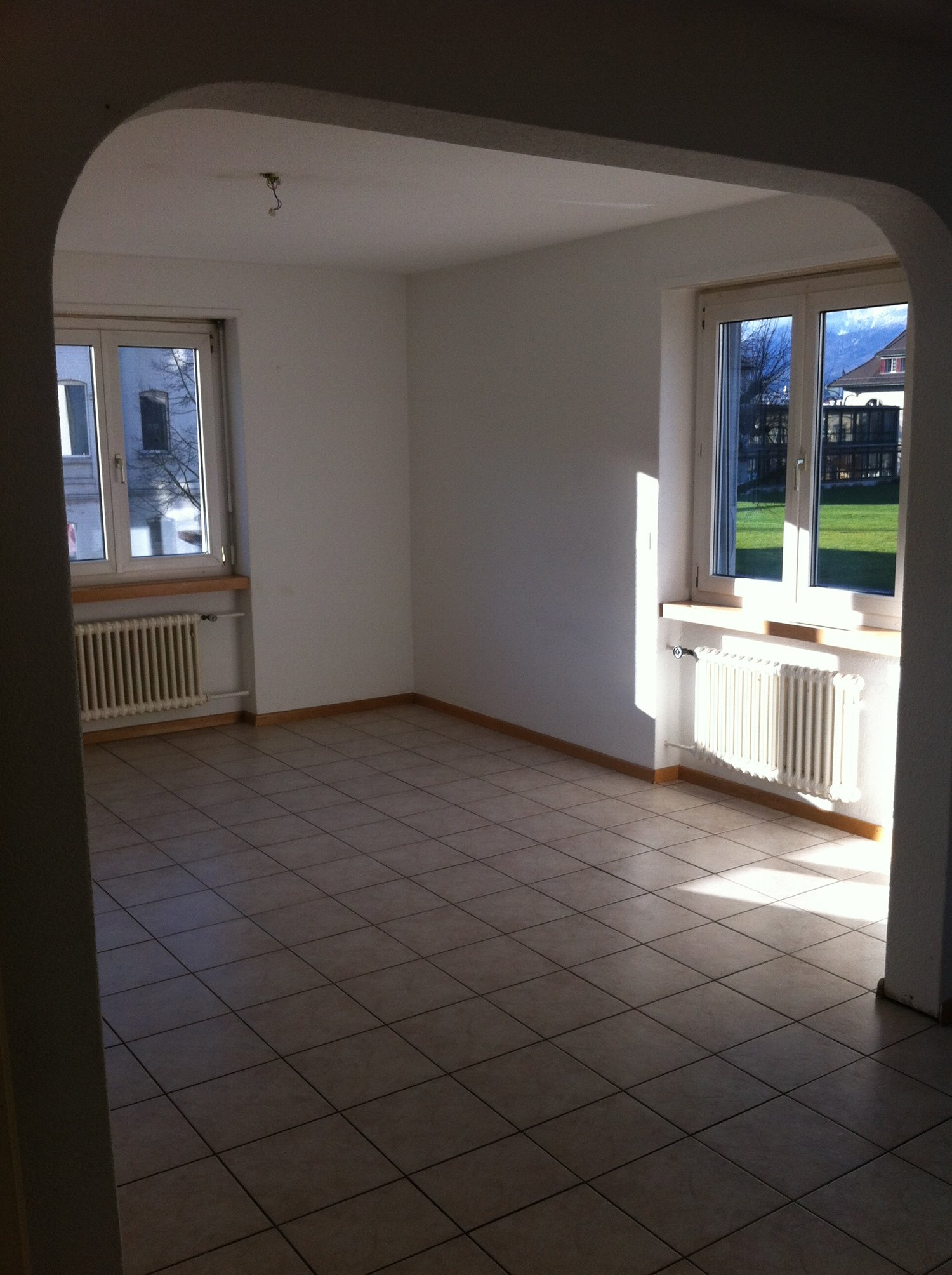 Spacious room with white walls, tiled flooring, and large windows overlooking a grassy outdoor area. The room appears to be empty, with no furniture or decor visible.