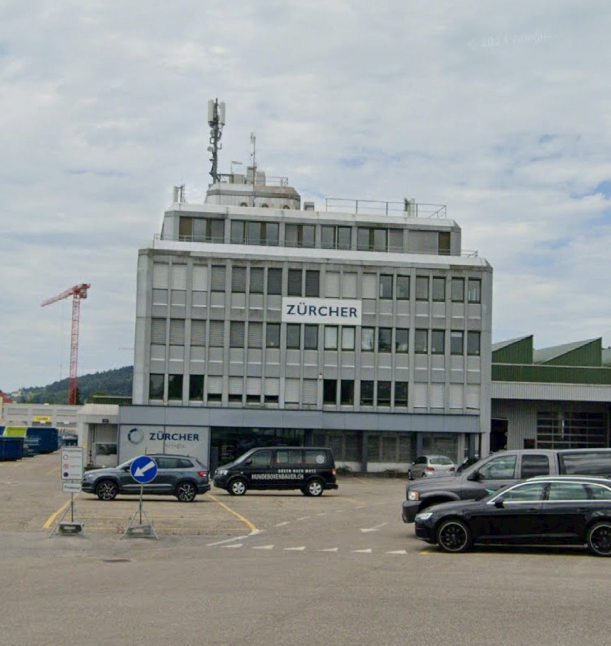 Multi-story office building with the Zurcher logo prominently displayed, surrounded by parked cars in a parking lot.