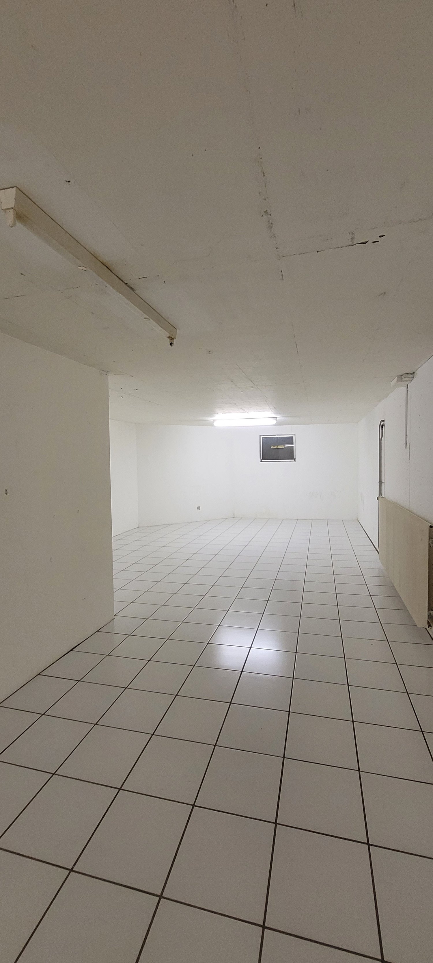 The image shows a large, empty room with white walls and a tiled floor. The room appears to be in a commercial or industrial building, with a simple light fixture visible on the ceiling.