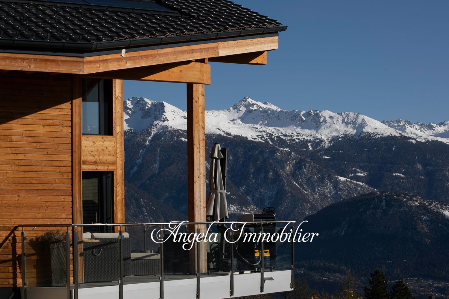 Wooden chalet-style building with a balcony overlooking snow-capped mountains in the background. The building has the name 'Angela Immobilier' displayed on the front.