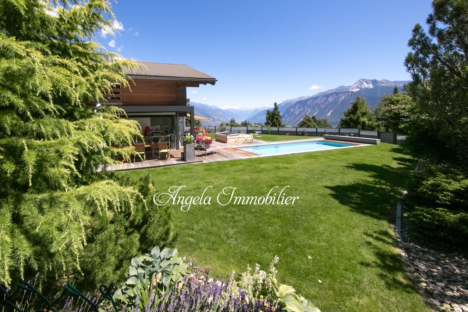 Luxury house with wooden exterior, surrounded by lush greenery and mountains in the background. The property features a swimming pool and offers a scenic view of the surrounding landscape.