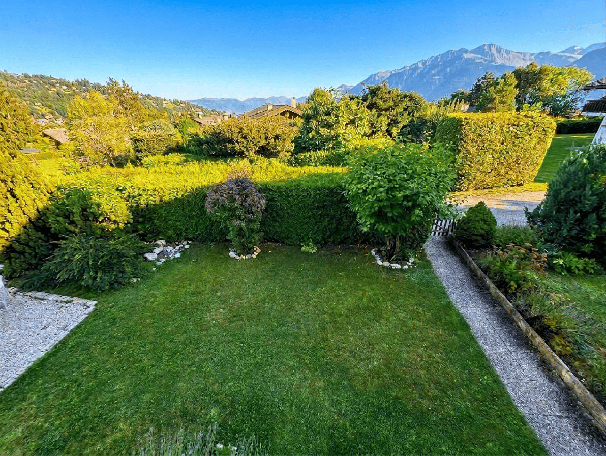 well-maintained garden with green lawn, hedges, and a gravel path, surrounded by trees and bushes, with a distant view of mountains
