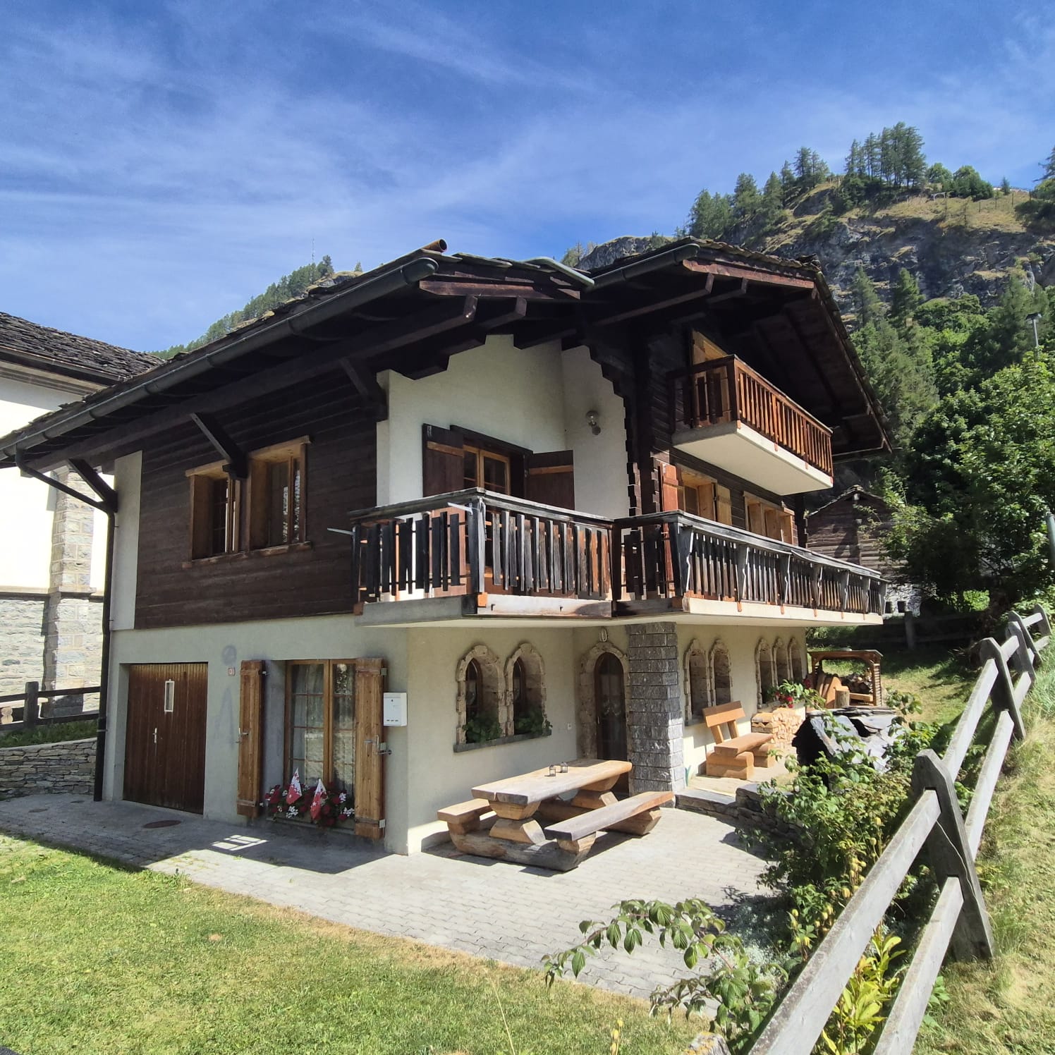 2-story wooden house, wooden balconies, white walls, wooden double doors, wooden picnic table, outdoor seating, wooden fence, grassy yard, mountainous landscape