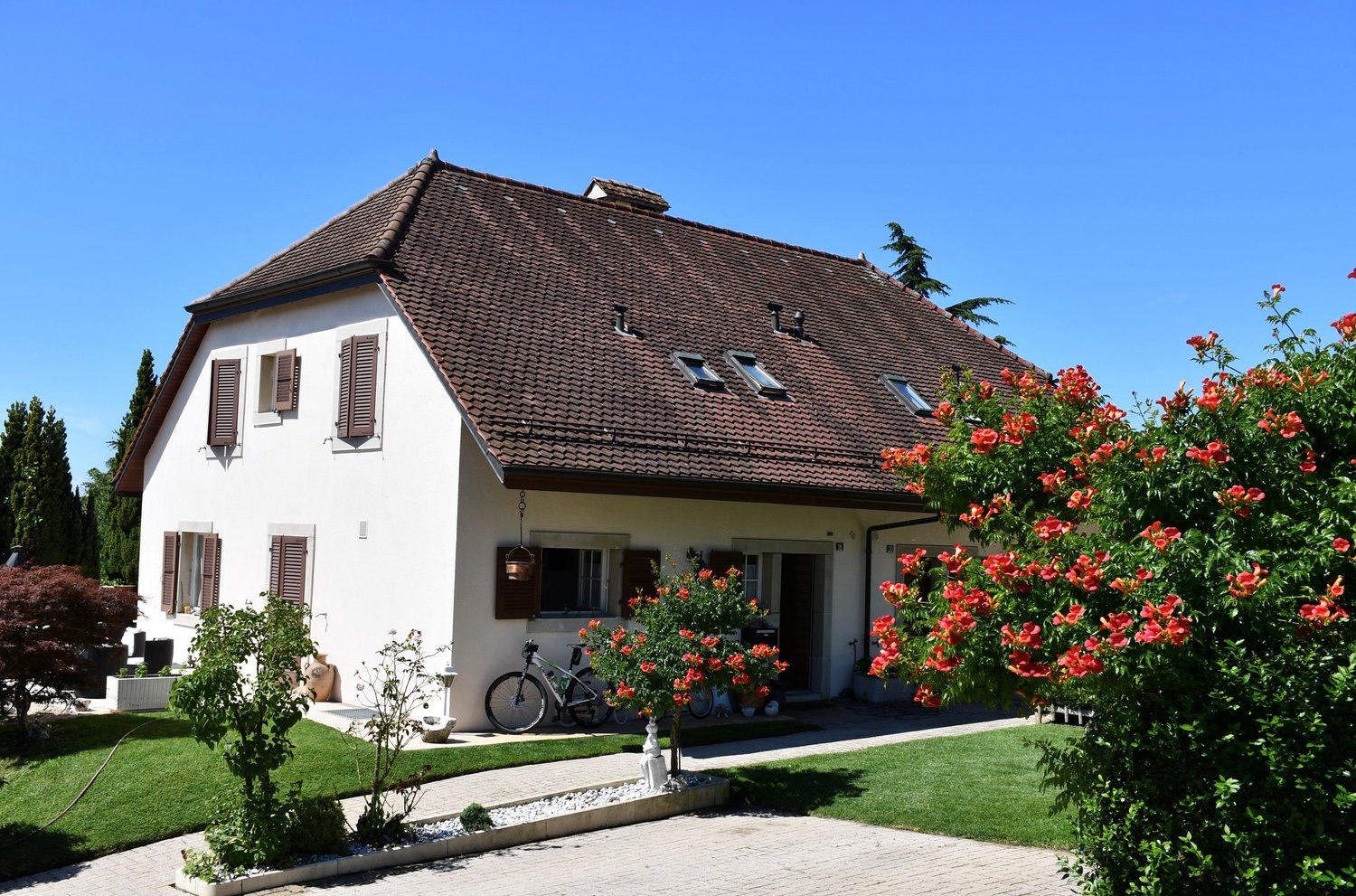2 story house, white exterior, brown roof, multiple windows, bicycle parked in front, garden with red flowers