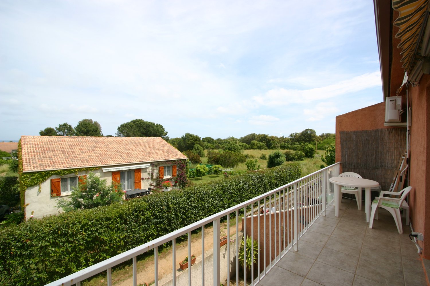 Two-story house with a tiled roof, surrounded by lush greenery and trees. The house has a balcony or terrace with a white railing, offering a scenic view of the surrounding countryside.