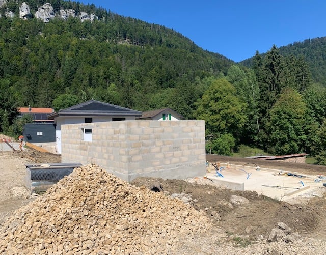 The image shows a house under construction in a mountainous, forested area. The foundation and walls of the house are visible, and there appears to be a garage or other structure next to the main building. The surrounding landscape is lush and green, with